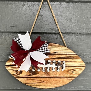 May include: A wooden football-shaped door hanger with a burgundy, white, and black checkered bow. The hanger is made of wood with a natural finish and has a rope hanger.