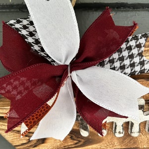 May include: A maroon, white, and black checkered bow with a brown and white patterned ribbon. The bow is tied around a wooden wreath with a football design.