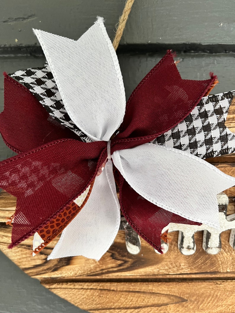 May include: A maroon, white, and black checkered bow with a brown and white patterned ribbon. The bow is tied around a wooden wreath with a football design.