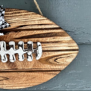 May include: A wooden football-shaped sign with a white painted football design. The sign is hanging from a twine string and has a black and white checkered bow.