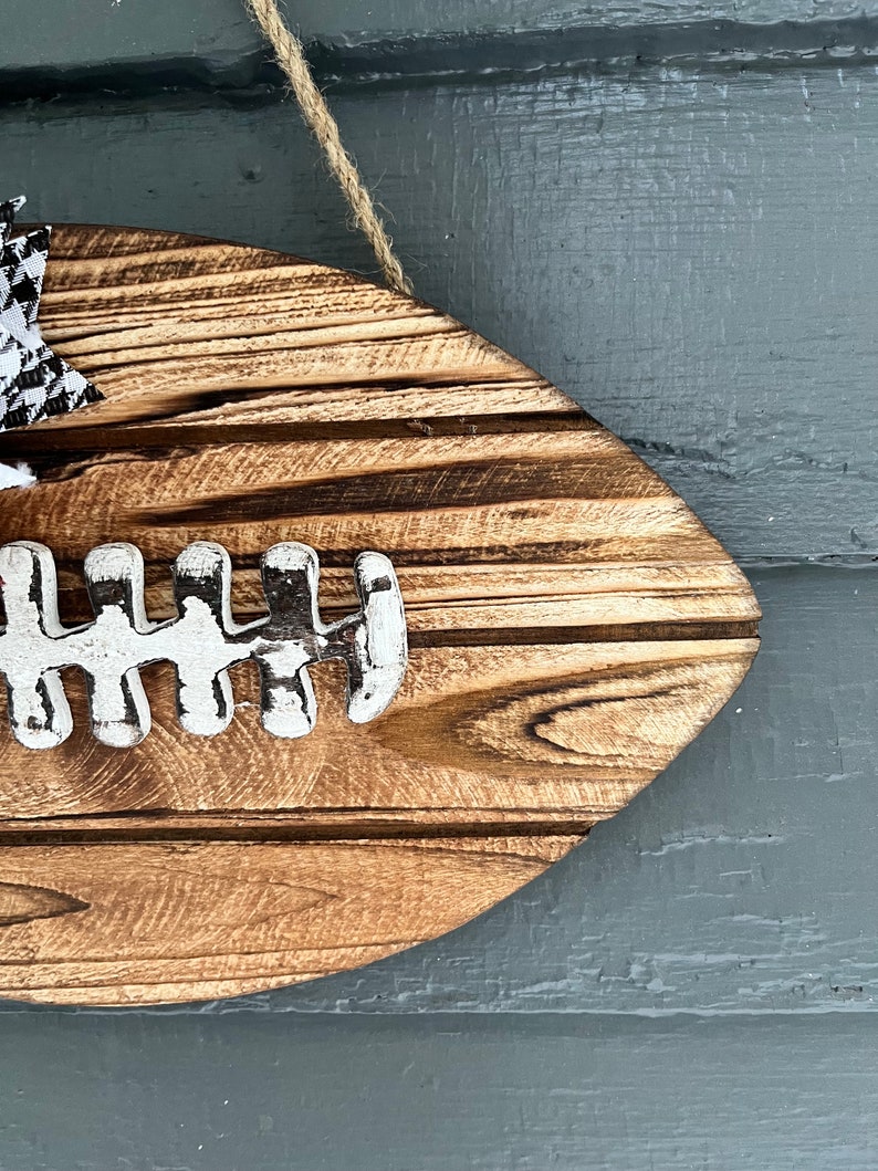May include: A wooden football-shaped sign with a white painted football design. The sign is hanging from a twine string and has a black and white checkered bow.