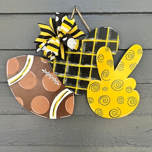May include: A decorative wooden wall hanging featuring a football, a heart with a plaid design, and a peace sign. The football is brown with white and yellow accents. The heart is black and yellow. The peace sign is yellow with black swirls.
