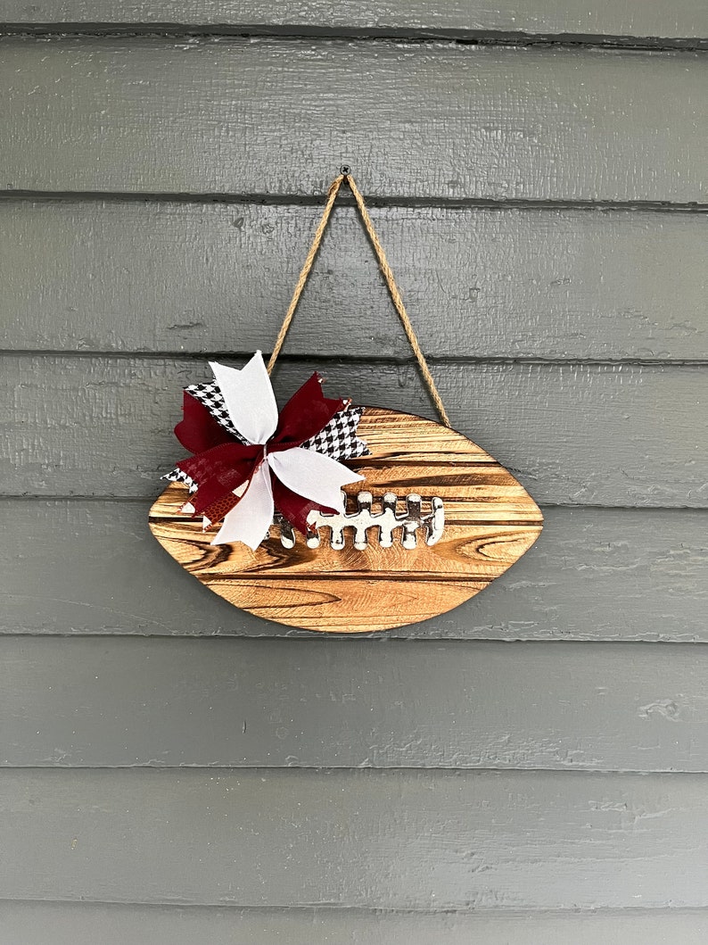 May include: A wooden football-shaped door hanger with a burgundy, white, and black checkered bow. The hanger is made of wood with a natural finish and has a rope hanger.