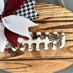 May include: A wooden football-shaped sign with a white painted football design and a maroon, white, and black bow.