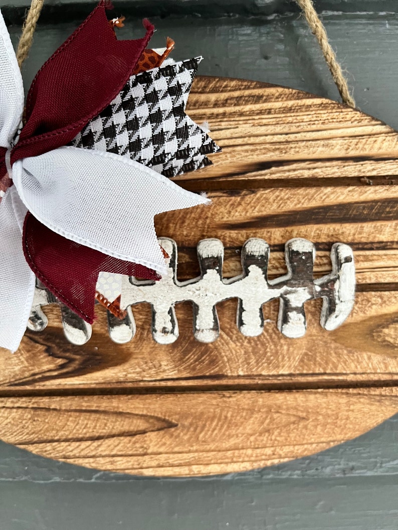 May include: A wooden football-shaped sign with a white painted football design and a maroon, white, and black bow.