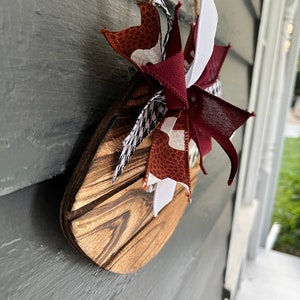 May include: A wooden sign with a brown, white, and red bow. The sign is shaped like a teardrop and has a wood grain pattern. The bow is made of fabric and has a white, brown, and red pattern.