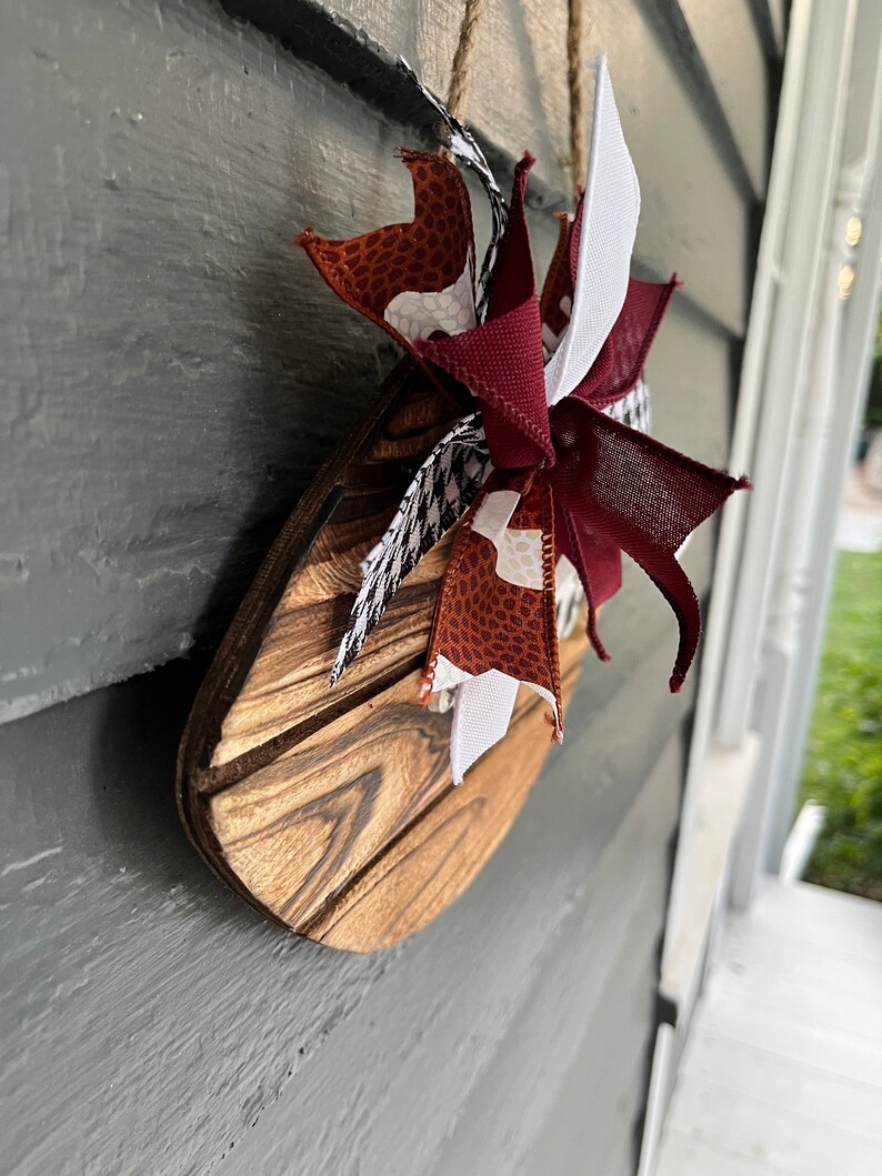 May include: A wooden sign with a brown, white, and red bow. The sign is shaped like a teardrop and has a wood grain pattern. The bow is made of fabric and has a white, brown, and red pattern.