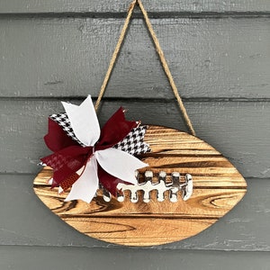 May include: A wooden football-shaped door hanger with a burgundy and white checkered bow. The football has a white painted lacing detail.