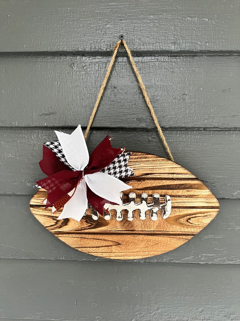 May include: A wooden football-shaped door hanger with a burgundy and white checkered bow. The football has a white painted lacing detail.