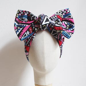 May include: A colorful headband with a large bow, displayed on a mannequin head. The bow features a vibrant geometric pattern in pink, black, red, blue, and white. The headband is a stylish accessory.