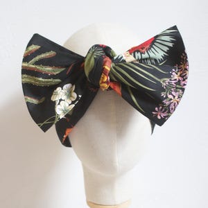 May include: A black fabric headband with a large bow, featuring a colorful floral and bird print. The bow is tied at the center and displayed on a mannequin head. The print includes cacti, flowers, and tropical birds.