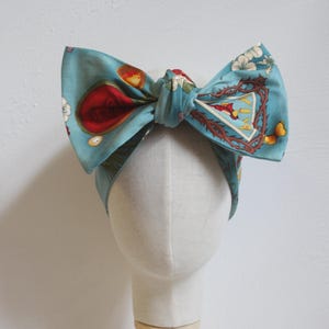 Op de afbeelding: Een turquoise stoffen hoofdband met een grote strik. De strik heeft een bloemen- en embleemdesign met rode, gele en witte accenten. De hoofdband wordt getoond op een mannequinhoofd met een houten standaard.