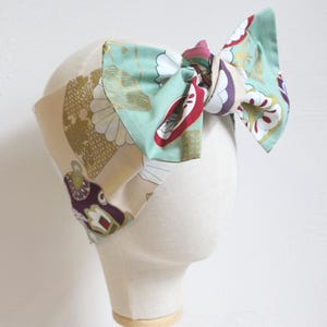 May include: A fabric headband with a large bow. The headband is beige with gold floral patterns. The bow is a mix of mint green, red, white, and purple floral patterns. The headband is displayed on a mannequin head.