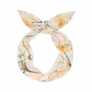 Könnte beinhalten: Ein cremefarbenes Stirnband mit floralem Muster und einer geknoteten Schleife. Der Stoff hat ein Design im Aquarellstil mit gelben und orangenen Blumen, grünen Stielen und grauer Schrift. Das Stirnband ist am Ansatz verdreht.