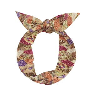 May include: A patterned headband with a knotted bow. The fabric features a repeating design of stylized waves and floral motifs in shades of brown, red, orange, and purple. The headband is designed to be worn around the head.