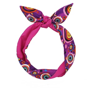 May include: A colourful headband with a retro design. The headband has a bright pink interior and a purple exterior decorated with colourful circular patterns in shades of orange, yellow, green, and blue. The headband is tied at the top in a bow.