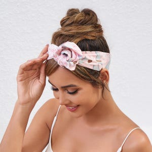 May include: A floral headband with a large rose-shaped knot. The headband features a pattern of pink and blue flowers on a light background. The woman in the image is wearing a white tank top.