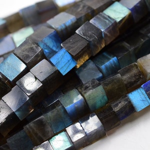 Puede incluir: Una hebra de cuentas de piedra preciosa de labradorita azul oscuro, cada cuenta es un cubo con un acabado facetado. Las cuentas tienen un brillo iridiscente con destellos de azul, verde y dorado.
