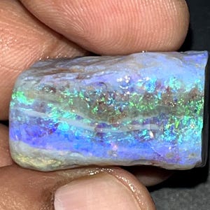 Könnte beinhalten: Ein roher, rechteckiger Opal-Edelstein mit einem lebendigen Farbspiel aus irisierenden Blitzen in Elektroblau, hellem Grün und violetten Nuancen. Der Stein weist eine texturierte, natürliche Oberfläche mit horizontalen weißen Mineraladern auf.