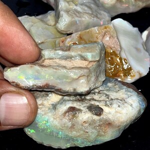 May include: A collection of raw, unpolished opal stones featuring irregular shapes and rough, textured surfaces. The stones display a milky white base color with vibrant flashes of green, yellow, and orange iridescence visible within the crystalline layers.