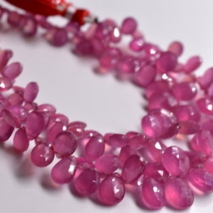 May include: A strand of pink gemstone beads, likely ruby, with a faceted, teardrop shape. The beads are strung on a red cord with silver accents.