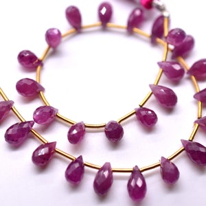May include: A strand of pink ruby gemstone beads in a teardrop shape. The beads are strung on a gold wire.