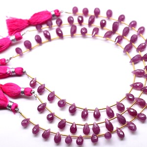 Op de afbeelding: Een streng roze edelsteen kralen, geregen op gouden draad, met roze kwastjes aan elk uiteinde. De kralen zijn gefacetteerd en hebben een druppelvorm.