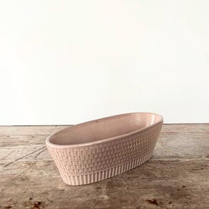 Puede incluir: Un macetero ovalado de cerámica de color rosa pálido con un diseño tejido texturizado. El macetero está sobre una superficie de madera.