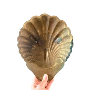 May include: A vintage, brass-toned seashell-shaped dish. The dish has a detailed, ribbed design and a scalloped edge. The brass has a patina with areas of green and dark spots, giving it a rustic appearance. The dish is held against a white background.