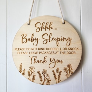 May include: A round wooden sign with the text "Shhh... Baby Sleeping" and "Please do not ring doorbell or knock. Please leave packages at the door. Thank you."  The sign is decorated with a floral border.