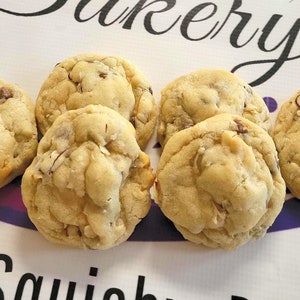 Op de afbeelding: Zes versgebakken chocolate chip cookies met zichtbare noten en chocoladestukjes. De koekjes zijn geplaatst voor een bord met de tekst "Sweet Squishy's Bakery" in een decoratief lettertype, met een paars en wit ontwerp.