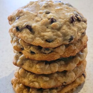 以下が含まれることがあります： 6つの黄金色のクッキーの山。クッキーはわずかに不均一な表面を持ち、濃い色のドライフルーツがちりばめられています。クッキーはオートミールレーズンクッキーのようです。
