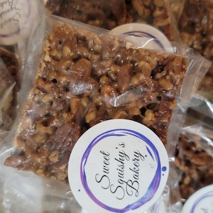 Könnte beinhalten: Mehrere einzeln verpackte Pekannuss-Kuchenriegel. Die Riegel sind rechteckig und mit Pekannüssen und einem karamellartigen Belag gefüllt. Jede Packung hat einen runden Aufkleber mit dem Text "Sweet Squishy's Bakery" in Lila und Weiß.