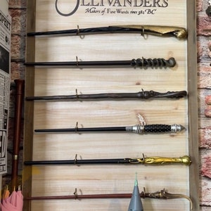 May include: A wooden sign with the text "Ollivanders Makers of Fine Wands since 382 BC" and a circle with a star inside. There are six wands hanging on the sign, each with a different design and color.