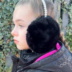 Puede incluir: Una persona joven que lleva una chaqueta negra con forro rosa y un gorro de orejas de piel sintética negra con ribete de perlas.