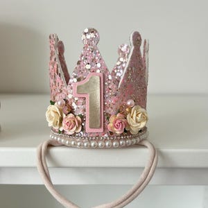 May include: A pink and gold glitter crown with the number 1, decorated with faux pearls and small fabric flowers. The crown is attached to a soft, elastic headband. A festive accessory for a first birthday celebration.