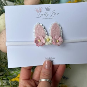 May include: A white headband with pink bunny ears, white lace trim, and small pink and white flowers. The card behind the headband reads "Dolly Bow Handmade Hair Bows" and has the Instagram handle "@dollybow".