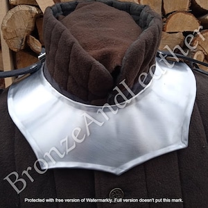 May include: A silver metal neck guard, also known as a gorget, worn over a brown quilted garment. The gorget is shaped like a crescent moon with a pointed bottom.