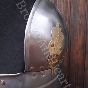 May include: A close-up of a silver metal breastplate with a gold leaf design. The breastplate has a leather strap attached to it.