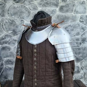 Peut inclure: Un gambeson rembourr&eacute; marron de style m&eacute;di&eacute;val avec une armure d'&eacute;paule en m&eacute;tal argent&eacute;. L'armure est rivet&eacute;e. Le gambeson a un col haut et est boutonn&eacute; sur le devant. Des lani&egrave;res en cuir fixent l'armure.