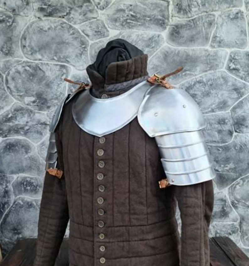 Peut inclure: Un gambeson rembourr&eacute; marron de style m&eacute;di&eacute;val avec une armure d'&eacute;paule en m&eacute;tal argent&eacute;. L'armure est rivet&eacute;e. Le gambeson a un col haut et est boutonn&eacute; sur le devant. Des lani&egrave;res en cuir fixent l'armure.