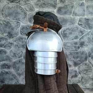 Peut inclure: Une &eacute;pauli&egrave;re en m&eacute;tal argent&eacute; avec un design en couches, fix&eacute;e par des sangles en cuir marron. L'&eacute;pauli&egrave;re est port&eacute;e sur un v&ecirc;tement rembourr&eacute; marron, sugg&eacute;rant une armure ou un costume m&eacute;di&eacute;val.