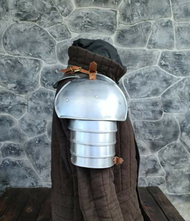 Peut inclure: Une &eacute;pauli&egrave;re en m&eacute;tal argent&eacute; avec un design en couches, fix&eacute;e par des sangles en cuir marron. L'&eacute;pauli&egrave;re est port&eacute;e sur un v&ecirc;tement rembourr&eacute; marron, sugg&eacute;rant une armure ou un costume m&eacute;di&eacute;val.