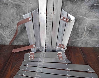 May include: A silver metal armor set with a chest plate and a separate set of plates for the legs. The armor is made of multiple plates that are connected with leather straps and rivets. The armor is laid out on a wooden surface.