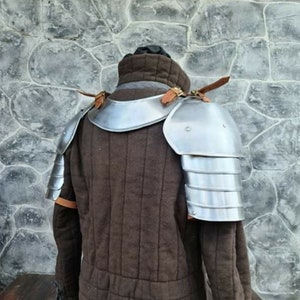 Peut inclure: Un gambison rembourr&eacute; marron de style m&eacute;di&eacute;val avec des &eacute;pauli&egrave;res en acier attach&eacute;es. L'armure est fix&eacute;e avec des lani&egrave;res de cuir. Le gambison a un col haut, pr&eacute;sent&eacute; devant un mur de pierre.