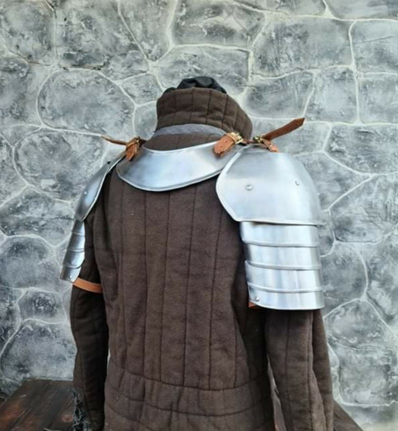 Peut inclure: Un gambison rembourr&eacute; marron de style m&eacute;di&eacute;val avec des &eacute;pauli&egrave;res en acier attach&eacute;es. L'armure est fix&eacute;e avec des lani&egrave;res de cuir. Le gambison a un col haut, pr&eacute;sent&eacute; devant un mur de pierre.