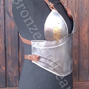 May include: A silver metal breastplate with a decorative gold design. The breastplate is worn on a mannequin with brown leather straps.