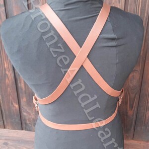 May include: A brown leather harness with straps that cross in the back. The harness has a buckle on the front and is worn on a black mannequin.