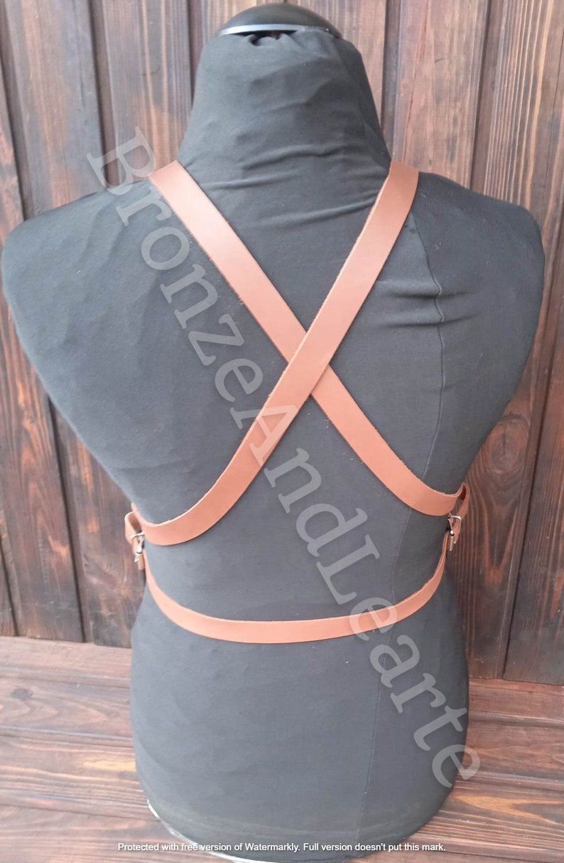 May include: A brown leather harness with straps that cross in the back. The harness has a buckle on the front and is worn on a black mannequin.
