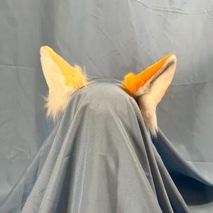 May include: A pair of fox ears on a headband. The ears are orange and white with a fluffy texture, and are attached to a thin, silver-colored metal headband. The ears are set against a blue background.